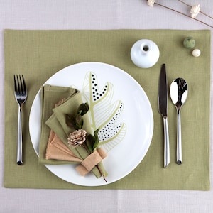 May include: A green placemat with a white plate featuring a stylized leaf design. A folded napkin with a decorative flower is placed on the plate. Silverware and a small white vase are also on the placemat.
