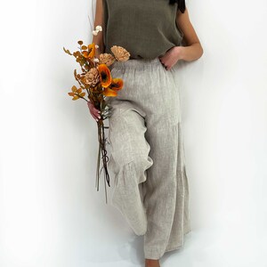 May include: An olive green sleeveless top with a round neckline paired with light beige wide-leg trousers featuring an elastic waistband and tiered hem detail. The outfit is shown alongside a bouquet of dried orange and cream-colored flowers.