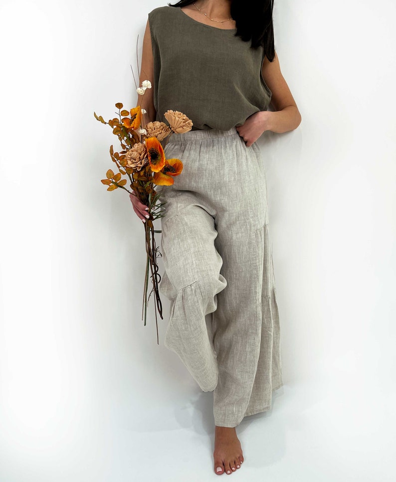 May include: An olive green sleeveless top with a round neckline paired with light beige wide-leg trousers featuring an elastic waistband and tiered hem detail. The outfit is shown alongside a bouquet of dried orange and cream-colored flowers.
