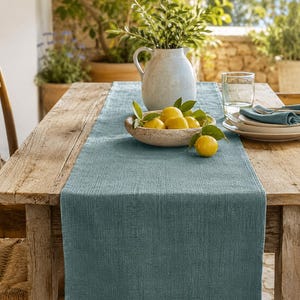 May include: A rectangular blue linen table runner with a visible woven texture draped over a rustic wooden table. The runner is centered beneath a ceramic bowl filled with bright yellow lemons and a white pitcher containing green leafy branches.
