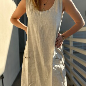 May include: A sleeveless beige dress featuring a round neckline and two large patch pockets on the front. The garment is crafted from a textured fabric with a visible weave, falling in a straight silhouette that reaches below the knee.