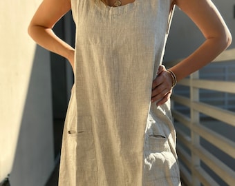 May include: A sleeveless beige dress featuring a round neckline and two large patch pockets on the front. The garment is crafted from a textured fabric with a visible weave, falling in a straight silhouette that reaches below the knee.