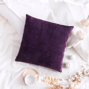 May include: A deep purple velvet pillow on a white bed with white decorative items and a cup of coffee on a saucer.