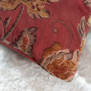 May include: A red throw pillow with a floral pattern in shades of brown and gold. The pillow has a zipper closure.