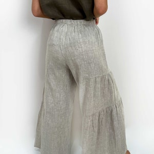 May include: Light gray, wide-leg trousers featuring a tiered design with two horizontal seams creating ruffled sections. The pants have an elasticized waistband and are crafted from a textured fabric with visible weave patterns, extending to the floor.