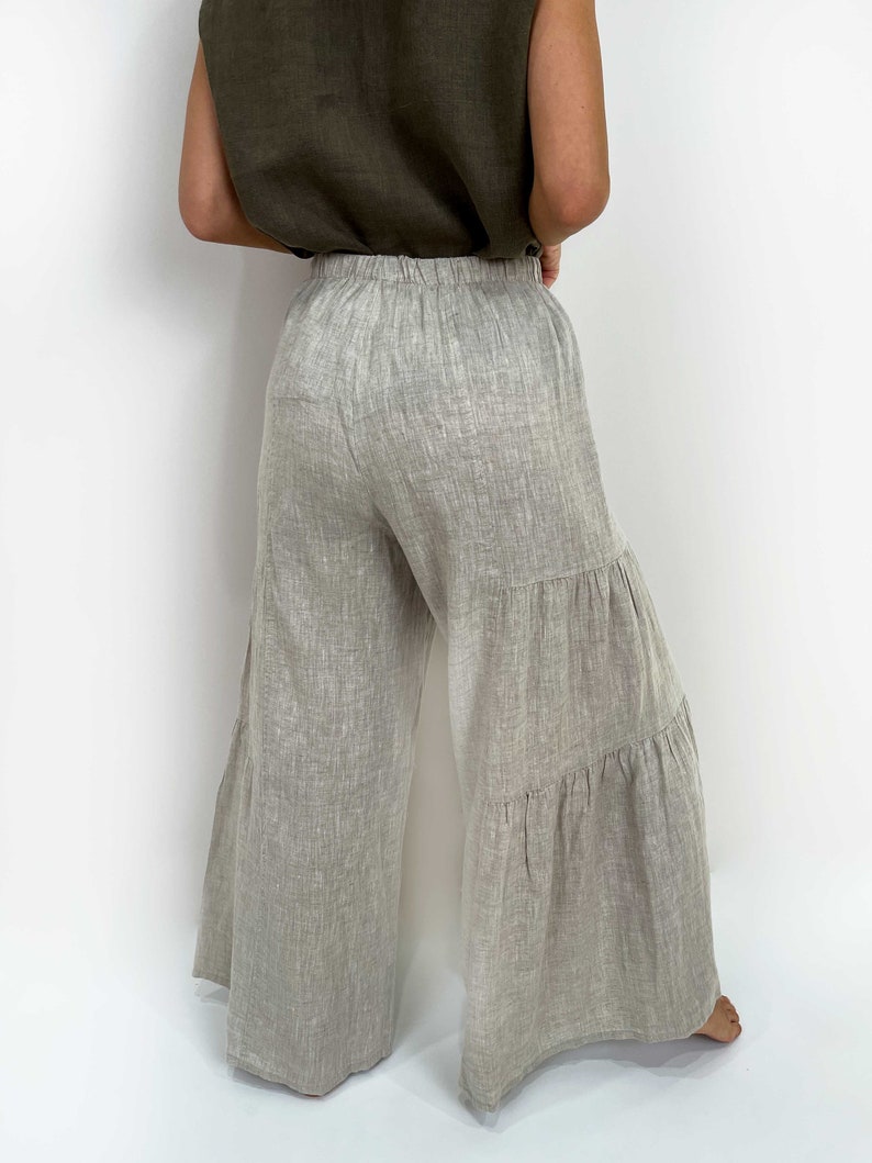 May include: Light gray, wide-leg trousers featuring a tiered design with two horizontal seams creating ruffled sections. The pants have an elasticized waistband and are crafted from a textured fabric with visible weave patterns, extending to the floor.