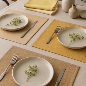 May include: A table setting featuring three rectangular placemats in shades of mustard, tan, and light brown. Each placemat holds a round, speckled off-white ceramic plate, a stainless steel fork or knife, and a small sprig of green foliage.