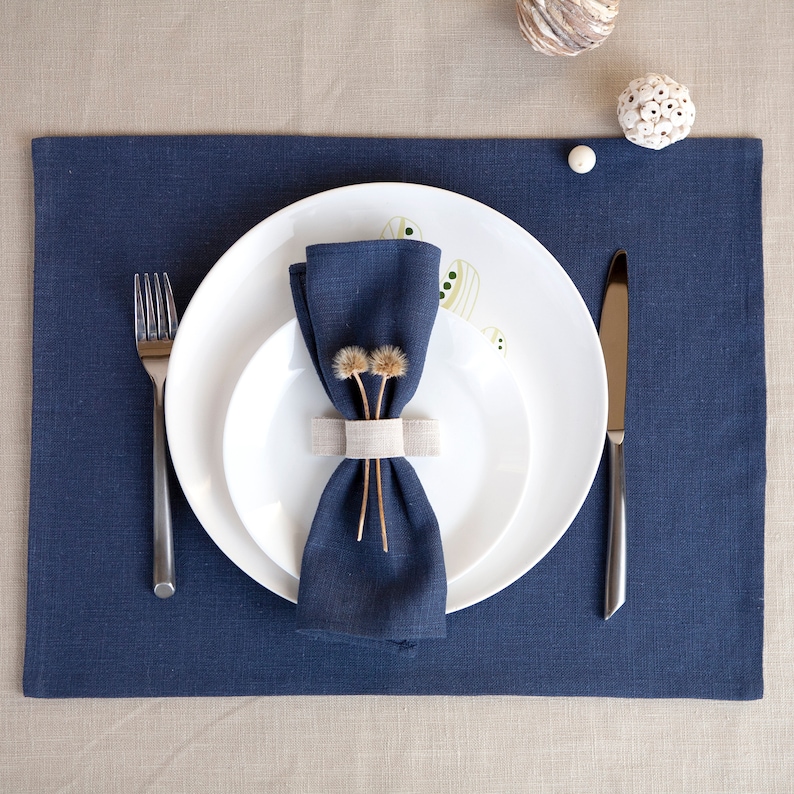 May include: A blue linen napkin is folded and placed on a white plate with a smaller white plate on top. The napkin is tied with a beige napkin ring and decorated with dried flowers. A silver fork and knife are placed on a blue linen placemat.