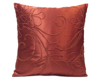 May include: A rust-colored throw pillow with an embroidered floral design. The pillow is made of a soft, silky fabric and has a square shape.