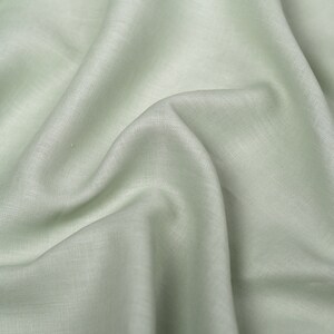 May include: A close-up of a light green fabric with a smooth, slightly textured surface. The fabric is folded and draped, creating soft folds and wrinkles.