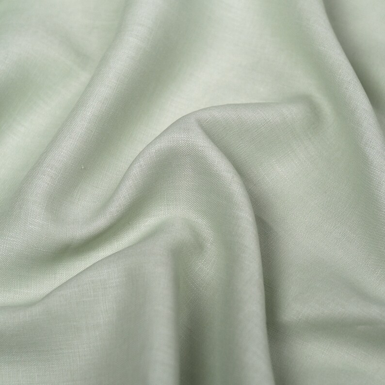 May include: A close-up of a light green fabric with a smooth, slightly textured surface. The fabric is folded and draped, creating soft folds and wrinkles.