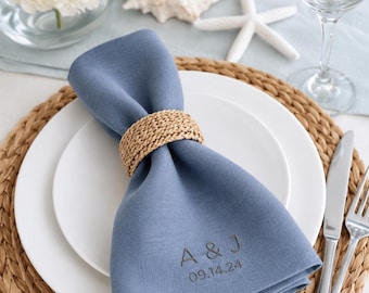 May include: A dusty blue fabric napkin secured with a woven tan rope napkin ring, resting on a white plate. The napkin is embroidered with the letters A & J and the date 09.14.24 in a dark thread, centred on the lower portion of the folded fabric.