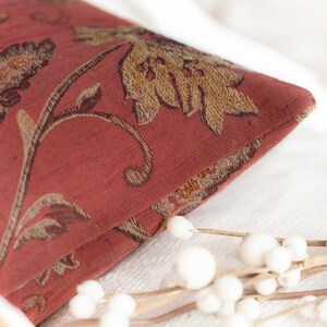 May include: Close-up of a red pillow with a floral pattern. The pillow has a brown and gold floral design with a textured fabric.