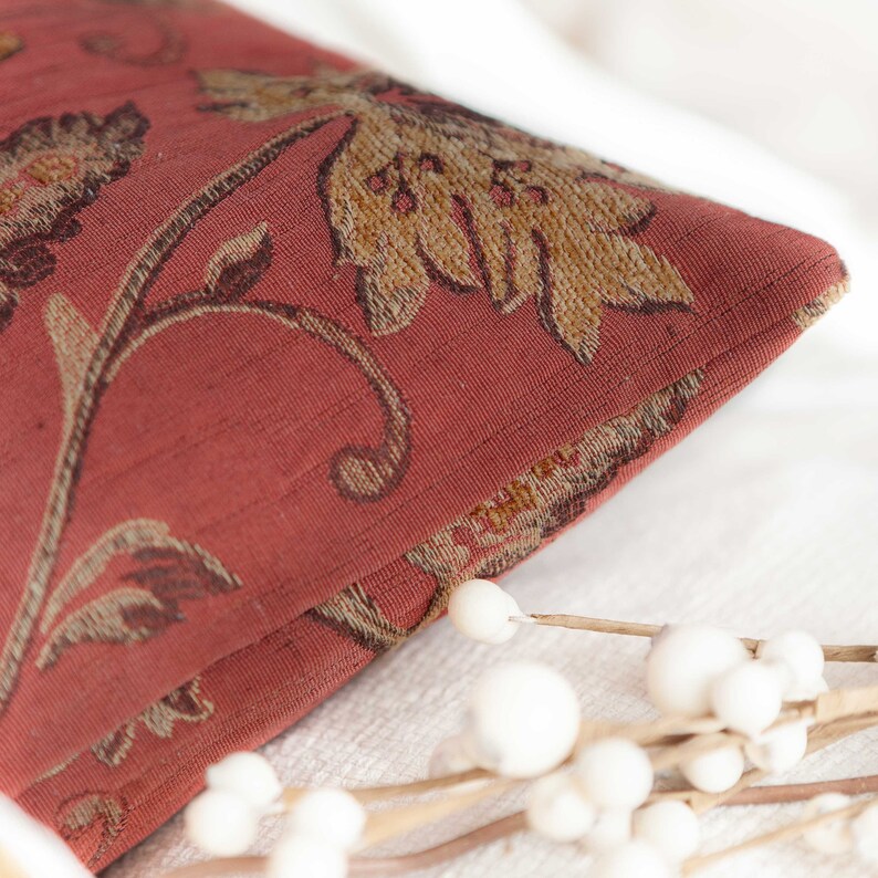 May include: Close-up of a red pillow with a floral pattern. The pillow has a brown and gold floral design with a textured fabric.