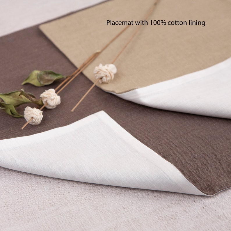 May include: A brown and beige placemat with a white cotton lining. The placemat is folded in half, showing the white lining. The placemat is on a light brown surface.