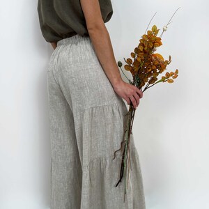 May include: Wide-leg trousers in a light beige, textured fabric featuring a tiered design with horizontal seams. The pants have an elasticized waistband and a loose, flowing silhouette. They are paired with a sleeveless olive green top.