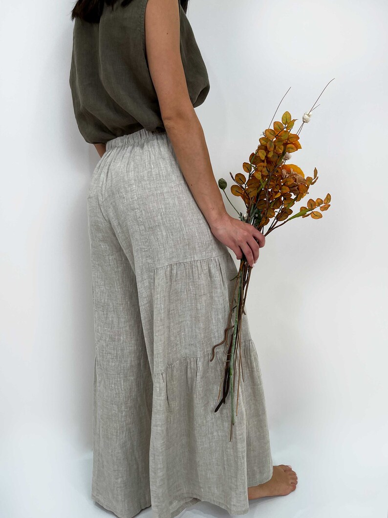 May include: Wide-leg trousers in a light beige, textured fabric featuring a tiered design with horizontal seams. The pants have an elasticized waistband and a loose, flowing silhouette. They are paired with a sleeveless olive green top.