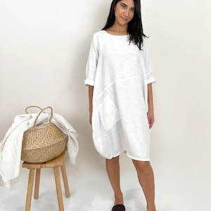 May include: A loose-fitting white knee-length dress with short sleeves, a round neckline, and a single patch pocket on the front. The garment features a visible horizontal seam across the midsection and a relaxed, flowing silhouette.