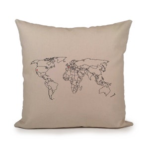 Puede incluir: Una almohada beige con un contorno negro de un mapa del mundo y dos corazones rojos que marcan ubicaciones en el mapa.