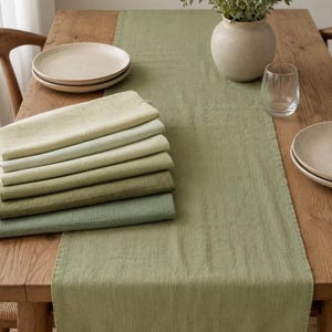 May include: A long sage green table runner made of textured fabric, accompanied by a stack of six folded napkins in a gradient of green shades, ranging from light pale green to a deeper, muted olive tone, all featuring a visible woven textile weave.