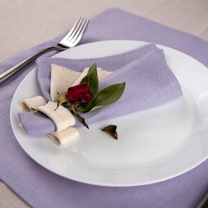 May include: A white ceramic dinner plate holds a folded lavender linen napkin secured with a cream-colored fabric napkin ring. A small dried dark red rosebud with green leaves rests on the napkin, and a silver fork lies to the side.