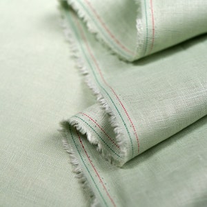May include: Close-up of a light green fabric with red and green stitching along the edge. The fabric has a subtle texture and a slightly frayed edge.