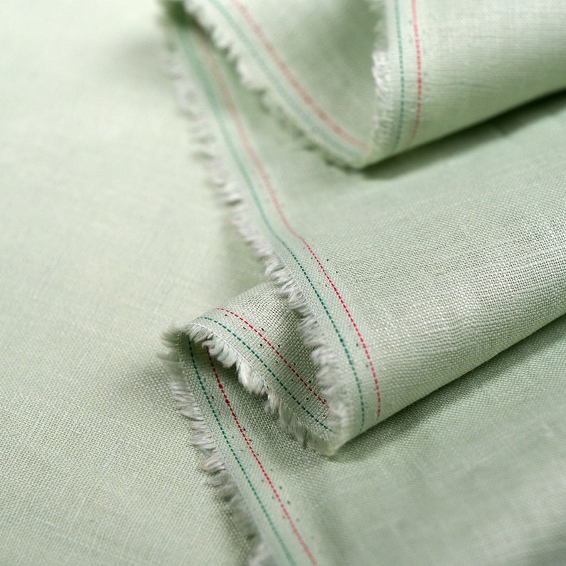 May include: Close-up of a light green fabric with red and green stitching along the edge. The fabric has a subtle texture and a slightly frayed edge.