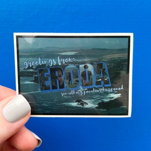 Könnte beinhalten: Ein Aufkleber mit einem blauen und weißen Design, auf dem "Greetings from Erodia something peculiar happened" steht. Der Aufkleber zeigt ein Bild eines Gewässers mit felsiger Küste und bewölktem Himmel.