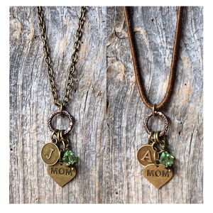 May include: Two necklaces with charms, one with a gold chain and the other with a brown cord. Both necklaces have a heart charm with the word "MOM" on it, a small round charm with the letter "J" on one and the letter "A" on the other, and a green crystal charm.