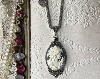 May include: A silver chain necklace with a cameo pendant featuring a white woman's profile. The cameo is set in a silver frame with black enamel accents. The necklace also has a small silver heart charm.