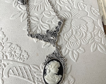 Op de afbeelding: Een zilveren ketting met een cameo-hanger met een wit silhouet van een vrouw op een zwarte achtergrond. De cameo is omgeven door een zilveren filigraanontwerp.