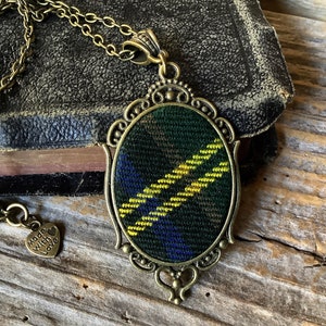 Puede incluir: Un collar de cadena dorada con un colgante que presenta un diseño de tela a cuadros verde, amarillo y azul en un marco ovalado adornado. El colgante tiene un dije en forma de corazón con el texto "Made with Love".