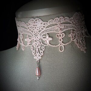 Puede incluir: Un delicado collar gargantilla de encaje rosa claro con intrincados patrones florales y de volutas. Un colgante de cuentas rosa en forma de pera cuelga del centro. El collar se exhibe en un maniqu&iacute;.