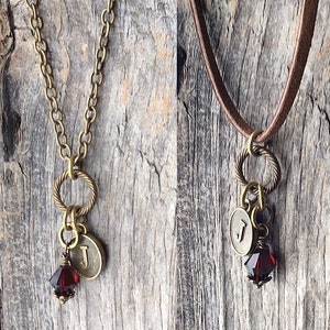 Peut inclure: Deux colliers avec un pendentif en forme de lettre 'J' et un pendentif en cristal rouge. Le collier de gauche a une chaîne dorée et le collier de droite a un cordon en cuir marron.