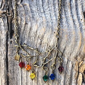 May include: A brass-toned necklace featuring a branch design with a leaf and six colorful crystal pendants. The pendants are red, orange, yellow, green, blue, and purple. The necklace hangs on a delicate chain.
