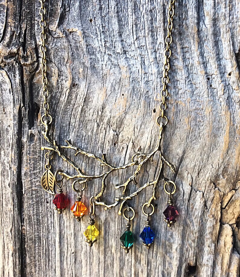 May include: A brass-toned necklace featuring a branch design with a leaf and six colorful crystal pendants. The pendants are red, orange, yellow, green, blue, and purple. The necklace hangs on a delicate chain.