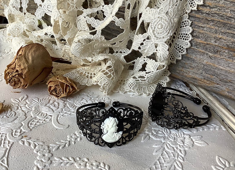 May include: Two black metal bracelets with intricate filigree designs. One bracelet features a white cameo with a woman's profile.