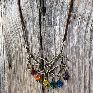 May include: A bronze-toned necklace featuring a branch design with colorful crystal pendants. The necklace has a brown suede cord and includes red, orange, yellow, green, blue, and purple crystal beads. A small leaf charm is also present.