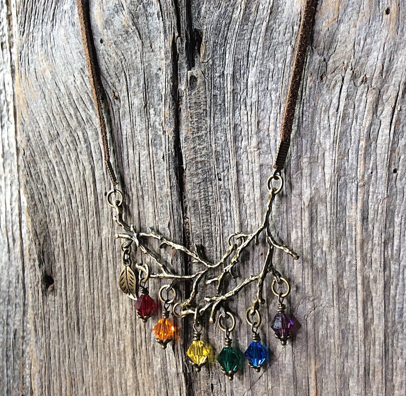 May include: A bronze-toned necklace featuring a branch design with colorful crystal pendants. The necklace has a brown suede cord and includes red, orange, yellow, green, blue, and purple crystal beads. A small leaf charm is also present.
