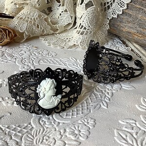 May include: Two black metal bracelets with intricate filigree designs. One bracelet features a white cameo with a woman's profile.