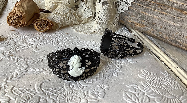 May include: Two black metal bracelets with intricate filigree designs. One bracelet features a white cameo with a woman's profile.