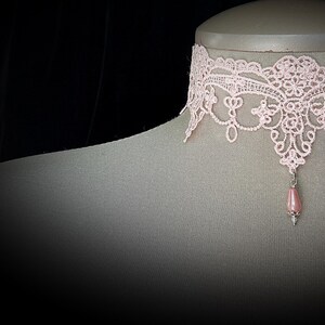 Puede incluir: Un collar gargantilla de encaje rosa claro con un colgante en forma de l&aacute;grima. El collar, expuesto sobre un maniqu&iacute;, presenta un intrincado dise&ntilde;o floral. El colgante es una cuenta rosa claro con una tapa plateada.