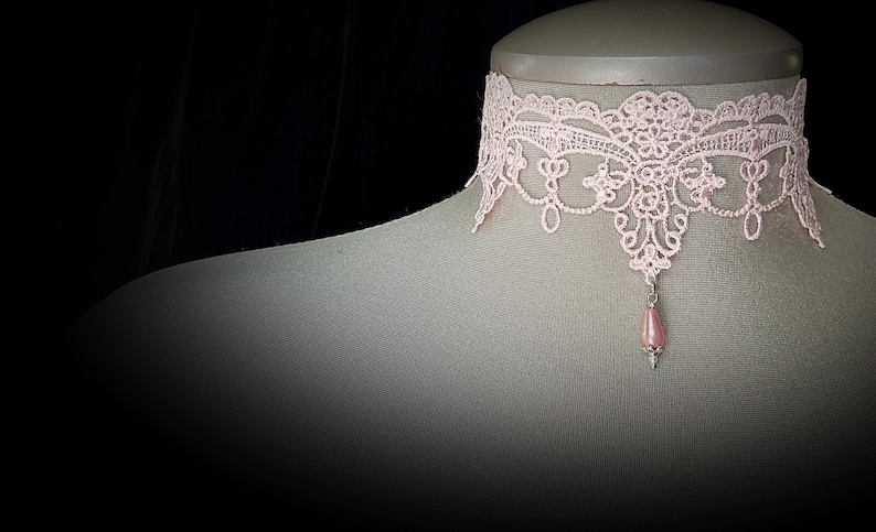 Puede incluir: Un collar gargantilla de encaje rosa claro con un colgante en forma de l&aacute;grima. El collar, expuesto sobre un maniqu&iacute;, presenta un intrincado dise&ntilde;o floral. El colgante es una cuenta rosa claro con una tapa plateada.