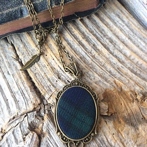 May include: A bronze chain necklace with a feather charm and an oval pendant featuring a green and blue plaid fabric design.