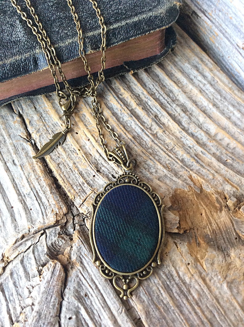 May include: A bronze chain necklace with a feather charm and an oval pendant featuring a green and blue plaid fabric design.