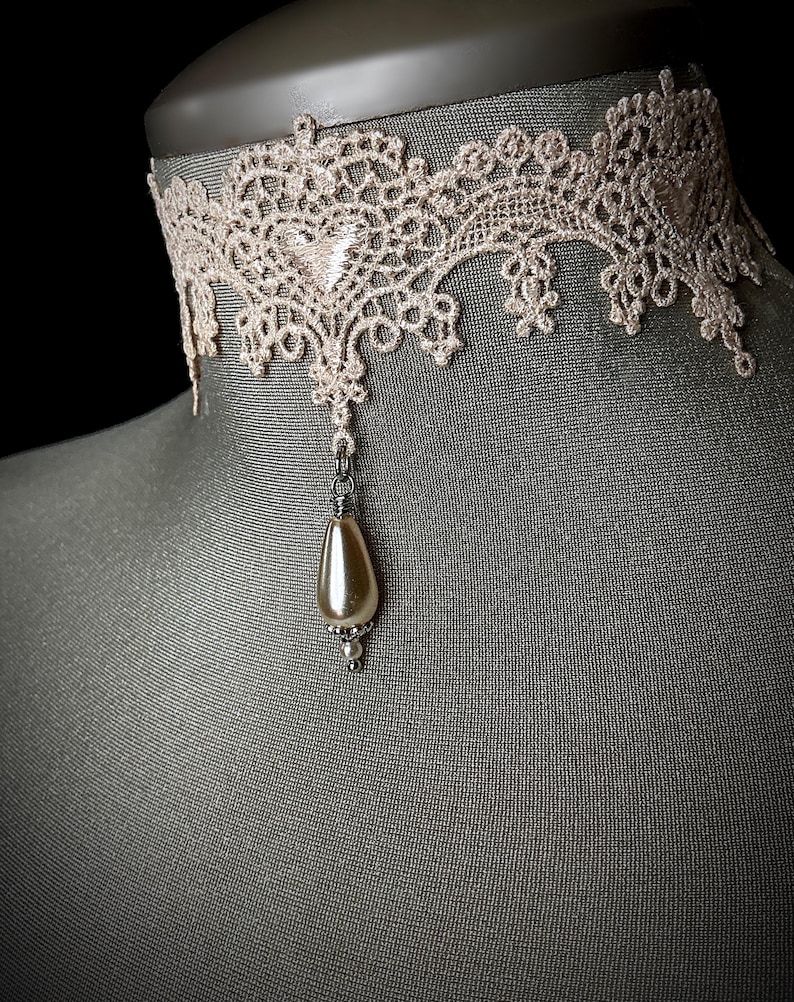 May include: A delicate ivory lace choker with a single pearl pendant. The choker is made of a fine, intricate lace pattern and is accented with a small, elegant pearl.