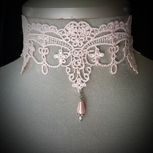 Puede incluir: Un delicado collar gargantilla de encaje rosa claro. El intrincado dise&ntilde;o de encaje presenta patrones florales y de volutas. Un peque&ntilde;o colgante de cuentas rosas en forma de pera cuelga del centro. El collar se exhibe en un maniqu&iacute;.