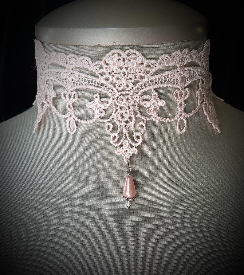 Puede incluir: Un delicado collar gargantilla de encaje rosa claro. El intrincado dise&ntilde;o de encaje presenta patrones florales y de volutas. Un peque&ntilde;o colgante de cuentas rosas en forma de pera cuelga del centro. El collar se exhibe en un maniqu&iacute;.