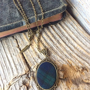 May include: A bronze chain necklace with a pendant featuring a dark green and blue fabric oval framed in an ornate bronze design. The necklace has a small feather charm.