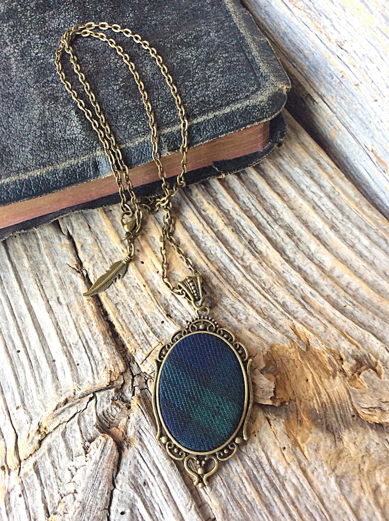 May include: A bronze chain necklace with a pendant featuring a dark green and blue fabric oval framed in an ornate bronze design. The necklace has a small feather charm.